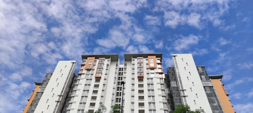 A high-rise residential building with modern architecture reaches towards a partly cloudy blue sky. The building features a combination of white and orange segments with numerous windows, and some parts have green foliage at the base.