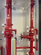 Two large red industrial pipes with pressure gauges and valves are mounted on a utility wall. The pipes have labels with numbers and are connected to other smaller pipes and devices.