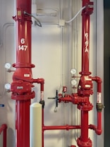 Two large red industrial pipes with pressure gauges and valves are mounted on a utility wall. The pipes have labels with numbers and are connected to other smaller pipes and devices.