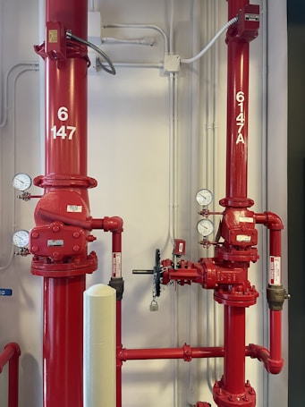 Two large red industrial pipes with pressure gauges and valves are mounted on a utility wall. The pipes have labels with numbers and are connected to other smaller pipes and devices.