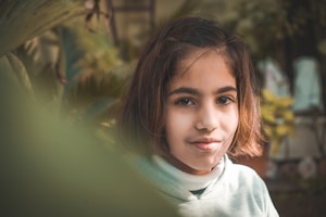 A young girl with shoulder-length hair is seen amidst a natural setting with greenery around. She is wearing a light-colored sweater and looking directly at the camera with a subtle smile.