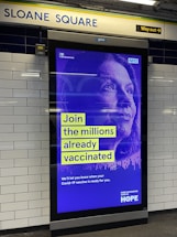 An advertisement poster located in a subway station with tiled walls. The poster has a blue and purple design featuring text that encourages joining the millions already vaccinated, with a pixelated image of a person's face in the background. The top corner displays government and NHS logos, indicating an official public health message.
