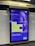 An advertisement poster located in a subway station with tiled walls. The poster has a blue and purple design featuring text that encourages joining the millions already vaccinated, with a pixelated image of a person's face in the background. The top corner displays government and NHS logos, indicating an official public health message.
