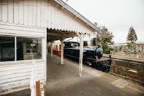 A vintage steam locomotive is stationed beside an old, wooden train platform. The surrounding architecture features rustic wooden siding and covered walkways. Trees and other vegetation are visible in the background, creating a nostalgic and historical ambiance.