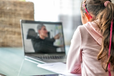 A young girl is sitting at a table, attentively looking at a laptop screen where a video call is taking place. She is wearing a pink hoodie with her hair tied back, and there is a notebook in front of her.