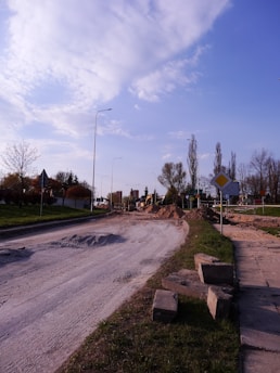 A partially constructed or under-construction road with loose gravel and sand piles. Construction machinery is visible in the background. The scene is set in a suburban area with street lamps and a mixture of leafless and budding trees along the roadside. A few traffic signs can also be observed.
