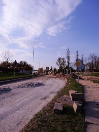 A partially constructed or under-construction road with loose gravel and sand piles. Construction machinery is visible in the background. The scene is set in a suburban area with street lamps and a mixture of leafless and budding trees along the roadside. A few traffic signs can also be observed.