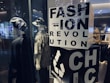 Two mannequins are dressed in casual, eclectic outfits, one wearing a hat and a dark cloak-like garment, while the other sports a bucket hat and a patterned shirt. A large block with bold letters spells out 'FASHION REVOLUTION CHIC' and includes a recycling symbol, suggesting a focus on sustainable fashion. Soft lighting and reflections from the glass create a modern, stylish ambiance.