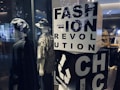 Two mannequins are dressed in casual, eclectic outfits, one wearing a hat and a dark cloak-like garment, while the other sports a bucket hat and a patterned shirt. A large block with bold letters spells out 'FASHION REVOLUTION CHIC' and includes a recycling symbol, suggesting a focus on sustainable fashion. Soft lighting and reflections from the glass create a modern, stylish ambiance.