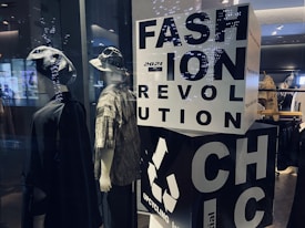 Two mannequins are dressed in casual, eclectic outfits, one wearing a hat and a dark cloak-like garment, while the other sports a bucket hat and a patterned shirt. A large block with bold letters spells out 'FASHION REVOLUTION CHIC' and includes a recycling symbol, suggesting a focus on sustainable fashion. Soft lighting and reflections from the glass create a modern, stylish ambiance.