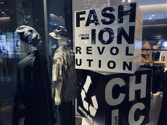 Two mannequins are dressed in casual, eclectic outfits, one wearing a hat and a dark cloak-like garment, while the other sports a bucket hat and a patterned shirt. A large block with bold letters spells out 'FASHION REVOLUTION CHIC' and includes a recycling symbol, suggesting a focus on sustainable fashion. Soft lighting and reflections from the glass create a modern, stylish ambiance.