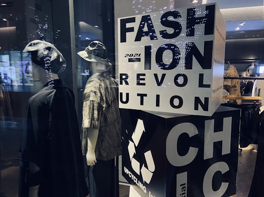 Two mannequins are dressed in casual, eclectic outfits, one wearing a hat and a dark cloak-like garment, while the other sports a bucket hat and a patterned shirt. A large block with bold letters spells out 'FASHION REVOLUTION CHIC' and includes a recycling symbol, suggesting a focus on sustainable fashion. Soft lighting and reflections from the glass create a modern, stylish ambiance.