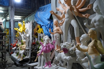 A workshop filled with unfinished statues, mostly of human figures in various poses. Some statues have multiple arms, and others are partially painted in vibrant colors such as yellow and pink. The environment appears cluttered, with a mix of finished and unfinished pieces. A large blue tarp hangs from the ceiling, and the lighting is dim with an artificial source overhead, creating shadows and a mysterious ambiance.