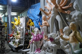 A workshop filled with unfinished statues, mostly of human figures in various poses. Some statues have multiple arms, and others are partially painted in vibrant colors such as yellow and pink. The environment appears cluttered, with a mix of finished and unfinished pieces. A large blue tarp hangs from the ceiling, and the lighting is dim with an artificial source overhead, creating shadows and a mysterious ambiance.