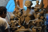 A sculptor works on a large clay statue depicting a multi-armed deity, surrounded by several smaller figures. The sculptures are in varying stages of completion, set against a rustic indoor backdrop with bluish light filtering through.