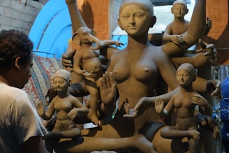 A sculptor works on a large clay statue depicting a multi-armed deity, surrounded by several smaller figures. The sculptures are in varying stages of completion, set against a rustic indoor backdrop with bluish light filtering through.