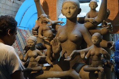 A sculptor works on a large clay statue depicting a multi-armed deity, surrounded by several smaller figures. The sculptures are in varying stages of completion, set against a rustic indoor backdrop with bluish light filtering through.