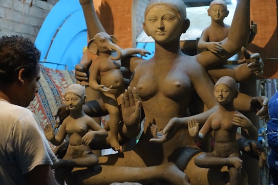 A sculptor works on a large clay statue depicting a multi-armed deity, surrounded by several smaller figures. The sculptures are in varying stages of completion, set against a rustic indoor backdrop with bluish light filtering through.