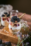 A glass jar is filled with layers of creamy white yogurt, crunchy granola, and vibrant red berries. Blackberries are artfully placed on top, and there is a golden spoon in the jar. The jar rests on a wooden surface with scattered seeds, surrounded by delicate purple and white flowers. The background is softly blurred, enhancing the focus on the jar.