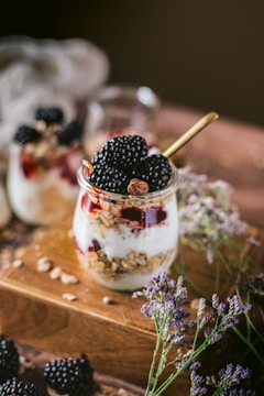 A glass jar is filled with layers of creamy white yogurt, crunchy granola, and vibrant red berries. Blackberries are artfully placed on top, and there is a golden spoon in the jar. The jar rests on a wooden surface with scattered seeds, surrounded by delicate purple and white flowers. The background is softly blurred, enhancing the focus on the jar.