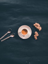 A cup of tea on a white saucer is placed on a dark surface next to two spoons and pieces of ginger root.
