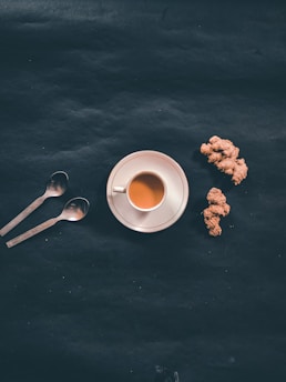 A cup of tea on a white saucer is placed on a dark surface next to two spoons and pieces of ginger root.