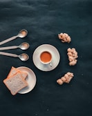 A neat arrangement of a cup of tea on a saucer sits beside slices of white bread on a plate. Three metallic teaspoons are aligned above the bread, and several pieces of ginger are scattered nearby, all set on a dark textured background.