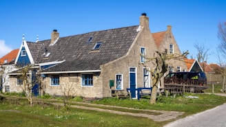 A charming rural scene featuring an old brick house with a steep, dark-tiled roof and bright blue windows. The house has a wooden door and several small trees in the front yard. A black van is parked on the gravel path beside the house. The sky is clear and blue, and a few small patches of grass and flowers are visible.
