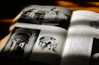 An open book under natural light, showing monochrome images of architecture and text. The left page displays architectural drawings and photographs, while the right page consists of textual content and a smaller image in the top right corner.