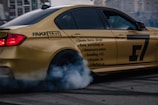 A golden sports car is performing a drift maneuver in an urban setting. Smoke billows from the rear tire, indicating high speed and tire friction. Various social media handles and branding are visible on the car's side. The background features blurred city buildings, suggesting motion.