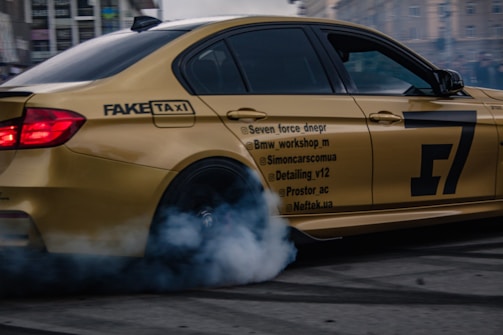 A golden sports car is performing a drift maneuver in an urban setting. Smoke billows from the rear tire, indicating high speed and tire friction. Various social media handles and branding are visible on the car's side. The background features blurred city buildings, suggesting motion.
