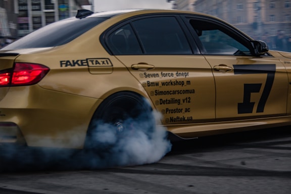 A golden sports car is performing a drift maneuver in an urban setting. Smoke billows from the rear tire, indicating high speed and tire friction. Various social media handles and branding are visible on the car's side. The background features blurred city buildings, suggesting motion.