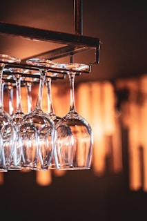A set of wine glasses hang upside down from a rack, reflecting warm ambient lighting. The background is softly blurred, highlighting the clarity and elegance of the glassware.