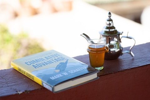 A book titled 'Cumulative Advantage' by Mark W. Schaefer lies on a wooden surface next to a small glass of tea and a traditional metal teapot. The scene appears to be set outdoors with a soft, blurry background.
