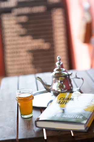 A wooden table holds a glass of amber-colored liquid, a silver teapot with a lid, a reflective tray, and a hardcover book titled 'Cumulative Advantage' by Mark W. Schaefer. The background features a blurred blackboard with white text, framed by a warm, soft focus ambiance.