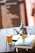 A wooden table holds a glass of amber-colored liquid, a silver teapot with a lid, a reflective tray, and a hardcover book titled 'Cumulative Advantage' by Mark W. Schaefer. The background features a blurred blackboard with white text, framed by a warm, soft focus ambiance.