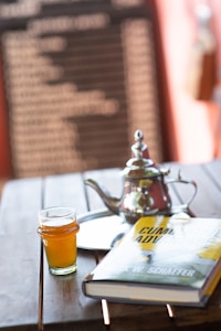 A wooden table holds a glass of amber-colored liquid, a silver teapot with a lid, a reflective tray, and a hardcover book titled 'Cumulative Advantage' by Mark W. Schaefer. The background features a blurred blackboard with white text, framed by a warm, soft focus ambiance.