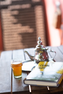 A wooden table holds a glass of amber-colored liquid, a silver teapot with a lid, a reflective tray, and a hardcover book titled 'Cumulative Advantage' by Mark W. Schaefer. The background features a blurred blackboard with white text, framed by a warm, soft focus ambiance.