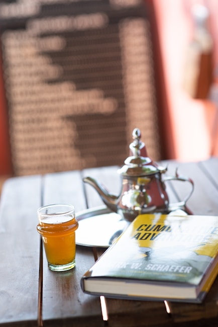 A wooden table holds a glass of amber-colored liquid, a silver teapot with a lid, a reflective tray, and a hardcover book titled 'Cumulative Advantage' by Mark W. Schaefer. The background features a blurred blackboard with white text, framed by a warm, soft focus ambiance.