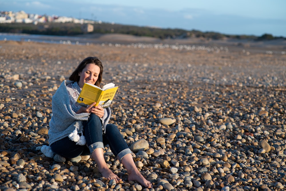 파란색 데님 재킷을 입은 여자가 낮 동안 책을 읽는 바위 해안에 앉아 있습니다.
