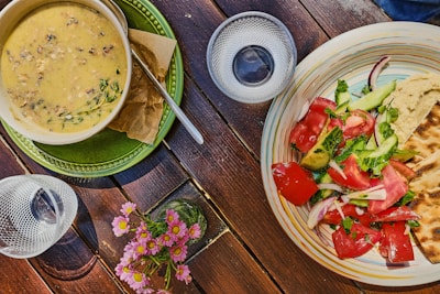 A rustic wooden table set with a vibrant plate of fresh salad including tomatoes, cucumbers, and herbs, accompanied by a portion of hummus and flatbread. Next to it is a hearty bowl of creamy soup garnished with herbs, placed on a green plate. There are empty glasses, and a small bouquet of pink flowers adds a touch of color to the setting.