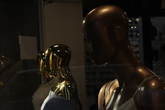 A display of mannequins with reflective golden surfaces in a dimly lit setting, creating a sleek and modern aesthetic. The foreground mannequin is bronze, while the others are gold, partially clothed, and arranged in proximity to each other, giving an impression of luxury and style.