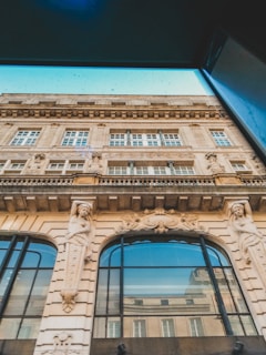 A detailed architectural facade of an old building, featuring large arched windows and ornate sculptures of human figures on either side. The facade is adorned with intricate carvings and decorations, emphasizing classical elements.