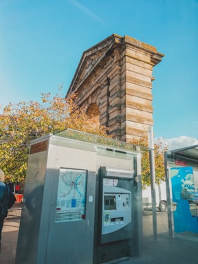 A ticket vending machine is positioned in the foreground, displaying transit maps and screens under a clear blue sky. Behind it, an ancient stone structure with classical elements looms, partially shaded by surrounding autumnal trees. The setup hints at a juxtaposition of modern transportation with historical architecture.