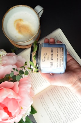 A hand holds a decorative candle with a rustic label amidst an open book, a cappuccino in a ceramic cup, and a bouquet of pink peonies and green foliage.