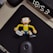 A cartoon character figurine is lying on a black mouse pad with its arms and legs splayed out. The character features large round glasses, a yellow shirt, blue shorts, and has a cheerful expression. The background includes an alarm clock showing the time 18:53 and a computer mouse placed below the figurine.