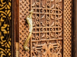 A detailed view of a wooden door with intricate carvings featuring geometric patterns and a prominent golden door handle shaped like a bird. The craftsmanship highlights traditional and ornate design elements.