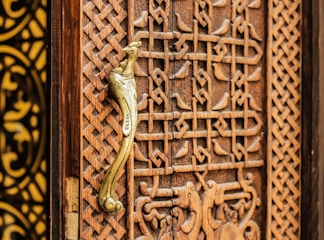 A detailed view of a wooden door with intricate carvings featuring geometric patterns and a prominent golden door handle shaped like a bird. The craftsmanship highlights traditional and ornate design elements.