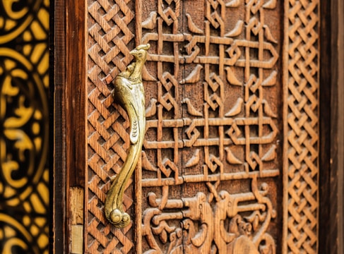 A detailed view of a wooden door with intricate carvings featuring geometric patterns and a prominent golden door handle shaped like a bird. The craftsmanship highlights traditional and ornate design elements.