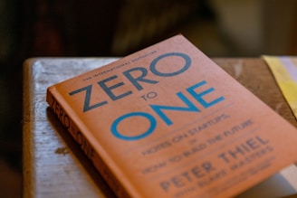 A book titled 'Zero to One' is resting on a wooden surface. The cover is orange with blue and white text, and the author is Peter Thiel. There is soft lighting, and the book's edges are slightly worn, suggesting it has been read or handled.